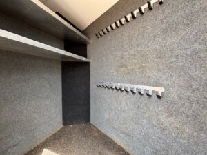 A small, carpeted storage room in the Cimarron 4 Horse trailer with metal shelves on the left wall and rows of metal hooks for hanging items on the right, perfect for organizing gear in your 15' Living Quarters. The floor is dark and textured. -Stillwater Trailer Sales