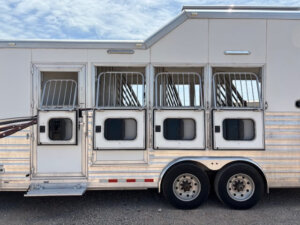A white Cimarron horse trailer with four open stall doors, each featuring a barred window and drop-down windows, is parked on gravel under a partly cloudy sky. This 4 horse side load model offers both style and functionality. -Stillwater Trailer Sales