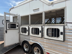 A Cimarron horse trailer in silver and white features three open side windows revealing stalls inside. The rear door is open, showing the entrance and interior bars. The trailer is parked outdoors under a blue sky. -Stillwater Trailer Sales