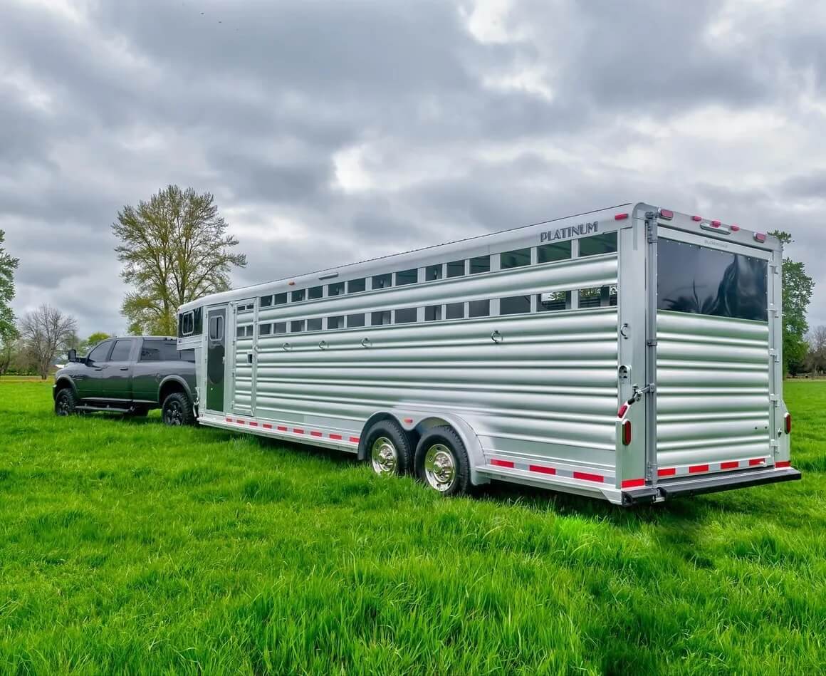 A large silver livestock trailer with ventilation slats is attached to a black pickup truck, parked on green grass under a cloudy sky. -Stillwater Trailer Sales