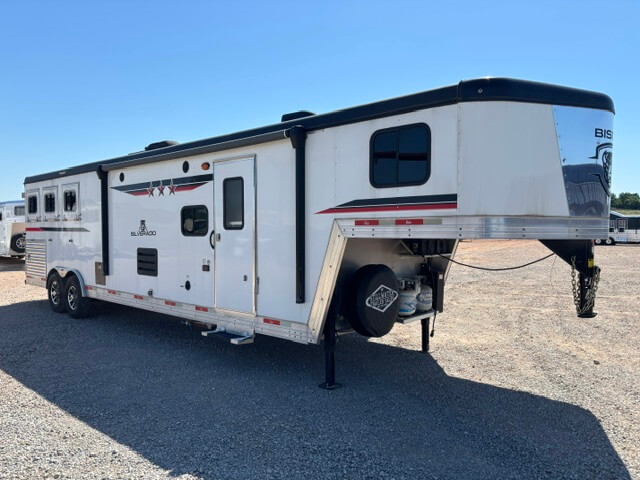 A large white gooseneck horse trailer with black and red accents is parked on gravel under a clear blue sky. The trailer has multiple windows and compartments and appears to have living quarters inside. -Stillwater Trailer Sales