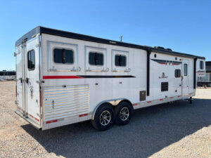A large white Bison 3 Horse trailer with living quarters, three windows, and two axles is parked on gravel. The trailer features exterior vents, red and black stripes, a side door with RV-style features, and sits under a clear blue sky. -Stillwater Trailer Sales