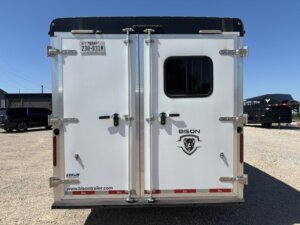 Rear view of a white Bison 3 Horse trailer with double doors, silver metal accents, a small window on the right door, and a Bison head logo below the window. Living Quarters and Texas license plate are also visible. -Stillwater Trailer Sales