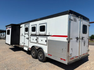 A large white 3 Horse Bison trailer with black trim and red accents is parked on gravel under a clear blue sky. The trailer features multiple windows, storage compartments, living quarters, and a slide-out section on one side. -Stillwater Trailer Sales