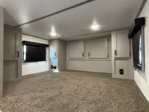 A spacious living quarters with carpeted flooring, gray walls, built-in cabinets, and two windows with black blinds, lit by ceiling lights. The space appears clean and minimalistic, ideal for a Bison 3 Horse setup. -Stillwater Trailer Sales