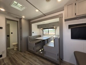Interior of a modern Bison 3 Horse RV living quarters with a dining area featuring two gray cushioned benches and a table, wood-look flooring, a small window with an outdoor view, and a visible bathroom door to the left. -Stillwater Trailer Sales