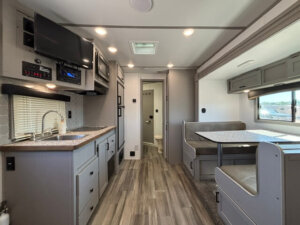 Interior of a Bison 3 Horse Living Quarters RV with a compact kitchen on the left&mdash;sink, stove, microwave, TV above. On the right is a dining booth with windows. Wood-style flooring throughout, and a door leads to the bathroom. -Stillwater Trailer Sales