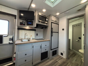 Modern Bison 3 Horse RV living quarters with sleek gray cabinets, sink, stove, microwave, TV, and refrigerator. The bathroom is visible through an open door, with wood-look flooring throughout. Bright, clean, and organized space. -Stillwater Trailer Sales