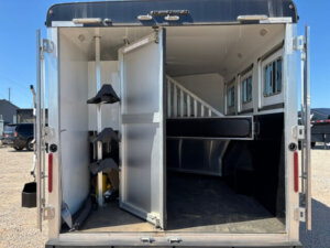 The image shows the open rear doors of a Bison 3 horse trailer, revealing interior stalls for horses and a tack storage area with mounted saddle racks on the left. The trailer is parked under a clear blue sky, hinting at comfortable horse living quarters inside. -Stillwater Trailer Sales