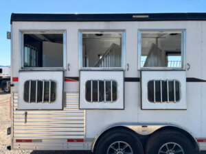 A white Bison 3 Horse trailer with three windows, each featuring metal bars. Two windows are open, revealing interior dividers, while the left window is closed. The Horse Living Quarters trailer sits on double wheels in the sun. -Stillwater Trailer Sales
