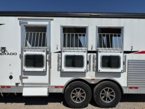 A white Bison 3 Horse trailer with living quarters features three barred windows&mdash;two open&mdash;and two wheels below. The trailer has a step and door on the left side and is parked on gravel under a clear blue sky. -Stillwater Trailer Sales