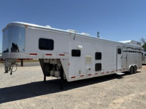 A large white gooseneck horse trailer with living quarters is parked on gravel under a clear blue sky. The trailer has multiple windows, side vents, and dual axles. -Stillwater Trailer Sales