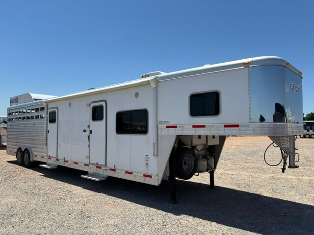 A large white gooseneck trailer with multiple windows and doors, parked on gravel under a clear blue sky. The trailer appears to have living quarters and space for transporting livestock. -Stillwater Trailer Sales