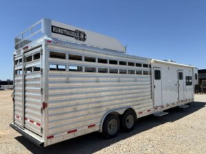 A large silver livestock trailer with ventilation slats, dual axles, and an attached white living quarters section, parked on gravel under a clear blue sky. -Stillwater Trailer Sales