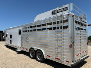 A large, silver livestock trailer with ventilation slats and dual rear axles is parked on gravel under a clear blue sky. The trailer has a rooftop rack and branding on an upper sign panel. -Stillwater Trailer Sales
