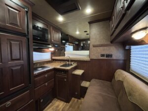 Interior of an RV with dark wood cabinetry, a small kitchen area with sink and stove, a sofa, and stairs leading to a raised sleeping area with a bed; overhead lights and window shades are visible. -Stillwater Trailer Sales