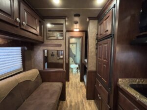 Interior of an RV featuring a brown sofa on the left, wooden cabinets and drawers, wood-look flooring, with a view into a small bathroom at the back containing a toilet and a shower. -Stillwater Trailer Sales