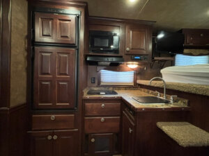 A compact RV kitchen with dark wood cabinets, a small sink, a stovetop, a microwave, a refrigerator, and a mounted TV. There is a window with blinds and a bunk bed visible to the right. -Stillwater Trailer Sales