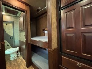 Small RV interior with dark wood paneling, featuring a compact bathroom with a toilet to the left, and two stacked bunk beds with gray bedding next to a large closet or cabinet on the right. -Stillwater Trailer Sales