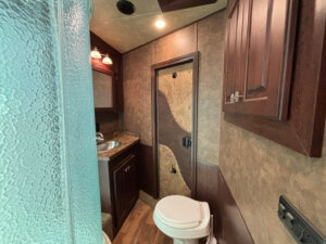 A compact RV bathroom with a glass shower on the left, a sink with a mirror and cabinet above, a toilet, wood cabinets, and a door with a decorative two-tone panel. The flooring is wood-style laminate. -Stillwater Trailer Sales