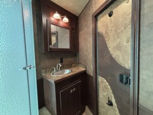 A compact bathroom with a small sink, brown wood cabinet, mirror with overhead lights, textured door, and part of a frosted glass shower visible on the left. -Stillwater Trailer Sales