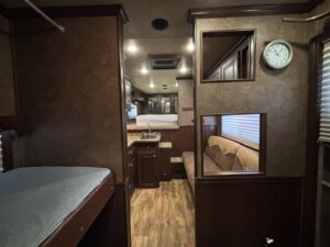 Interior of an RV with wood-paneled walls, a bunk bed on the left, small kitchen with sink and cabinets in the center, a beige couch on the right, and a wall clock above the couch. Light streams through window blinds. -Stillwater Trailer Sales
