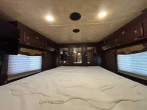 A cozy RV bedroom features a large bed with a white mattress, wooden cabinets above, mirrored closet doors at the foot of the bed, and windows with pleated shades on both sides. Warm ceiling lights illuminate the space. -Stillwater Trailer Sales
