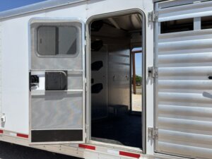 A close-up of an open door on a silver livestock trailer, revealing an interior area with several black saddle racks mounted on the wall. Sunlight and gravel are visible outside. -Stillwater Trailer Sales