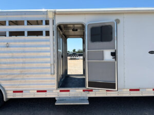 A white livestock trailer with an open door, revealing the interior. The trailer has horizontal slats on the left and a solid-panel section on the right. The ground is gravel, and other trailers are visible in the background. -Stillwater Trailer Sales