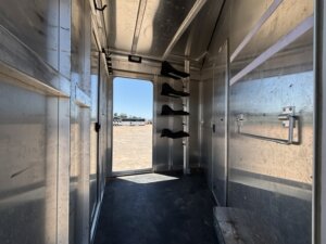 Interior of an empty metallic horse trailer with saddle racks on the right, open door at the end showing a sunny outdoor area, and hooks on the wall for equipment. -Stillwater Trailer Sales