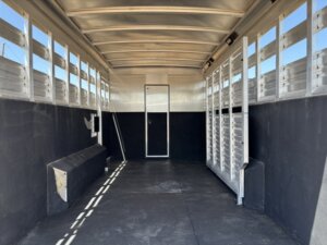 Interior of an empty livestock trailer with metal walls, slatted windows for ventilation, a small bench on the left, and a door at the far end; sunlight creates shadows on the floor. -Stillwater Trailer Sales