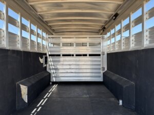 Interior view of an empty livestock trailer with metal slatted walls, black padded sides, and a closed central gate separating the front and back sections. Sunlight filters through the side slats. -Stillwater Trailer Sales