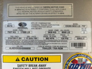 A close-up of a trailer information label showing manufacturer, model (Exiss 8032 LQ - 76), weight ratings, tire specs, VIN, and a yellow caution sticker for safety break-away requirements. -Stillwater Trailer Sales