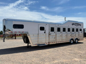 A large, white and silver gooseneck horse trailer with multiple doors and windows is parked on gravel under a blue sky. The trailer has Bloomer Trailers branding on the side. -Stillwater Trailer Sales