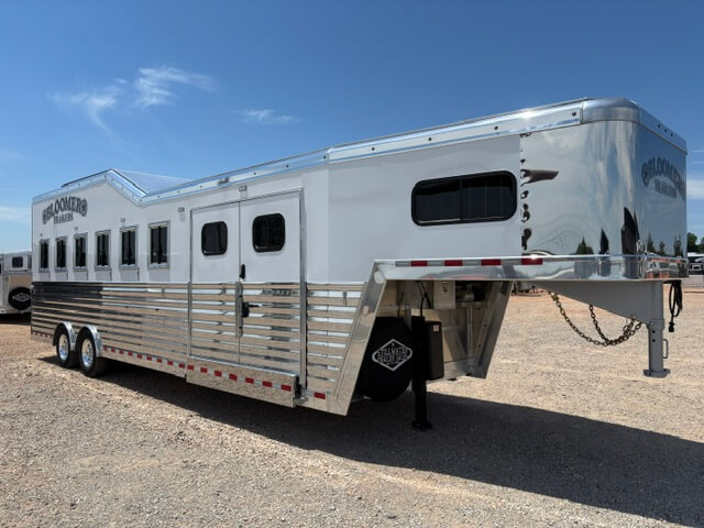 A large, silver gooseneck horse trailer with three axles and multiple windows is parked on gravel under a clear blue sky. The trailer has shiny metal siding and “Bloomer” branding visible on the front. -Stillwater Trailer Sales