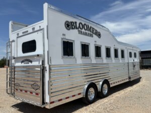 A large white and silver Bloomer horse trailer with multiple windows, shiny metal siding, dual axles, and a ramp, parked on gravel under a partly cloudy sky. -Stillwater Trailer Sales