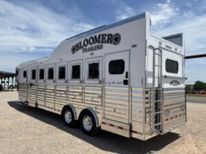 A large silver Bloomer horse trailer with multiple windows and vents is parked on gravel under a blue sky. The trailer has two axles, shiny metal siding, and a rear ladder. -Stillwater Trailer Sales
