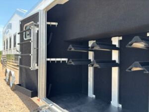 Open compartment of a horse trailer showing multiple black saddle racks and bridle hooks inside, with the trailer door open and sunlight illuminating the interior. -Stillwater Trailer Sales