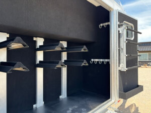 The image shows the inside of a horse trailer tack room with multiple black saddle racks on the left and rear walls, several hooks for hanging equipment, and a small window on the door. -Stillwater Trailer Sales
