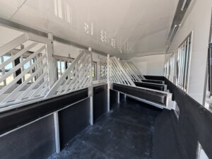 Interior of a trailer with metal dividers forming stalls, likely for transporting horses or livestock. The space is clean with black flooring, white walls, and barred windows along the sides. -Stillwater Trailer Sales