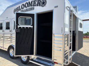 A silver Bloomer horse trailer is parked outdoors with a side door open, revealing a carpeted interior with shelves. The trailer has multiple windows and shiny metal accents under a bright blue sky. -Stillwater Trailer Sales
