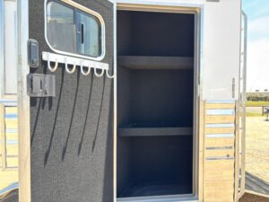 Open compartment door on a metallic trailer reveals two empty shelves inside; the door has hooks and a window. Sunlight and an outdoor setting are visible in the background. -Stillwater Trailer Sales