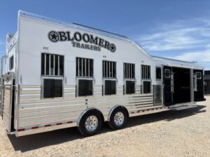 Large white Bloomer horse trailer with multiple barred windows and double rear doors, parked on gravel under a blue sky. -Stillwater Trailer Sales