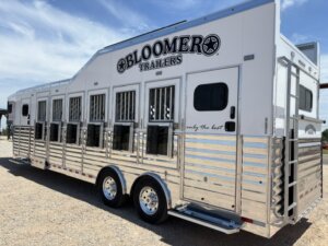 A large white and silver Bloomer Trailers horse trailer with multiple windows and vents, parked on gravel under a blue sky. -Stillwater Trailer Sales