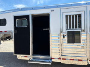 A silver horse trailer with an open side door, revealing a dark interior and saddle rack inside. The trailer has a barred window and shiny metallic exterior panels, parked on gravel under a blue sky. -Stillwater Trailer Sales