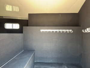 A clean, empty room with dark walls, a bench on the left, two small rectangular windows, and a white wall-mounted rack with multiple hooks on the right wall. -Stillwater Trailer Sales