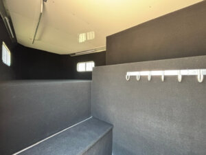 Interior of a trailer with black padded walls and benches, small barred windows, metal hooks on one wall, and a stepped platform area, likely designed for transporting animals such as horses. -Stillwater Trailer Sales