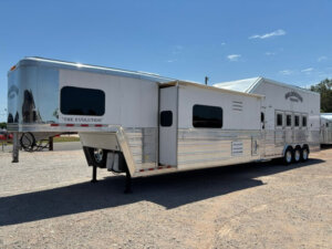 A large, silver Bloomer 4 Horse Trailer with a side load, multiple windows, and a slide-out section sits parked on gravel under a clear blue sky. THE EVOLUTION is labeled on the front. -Stillwater Trailer Sales
