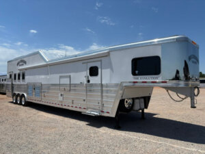 A large silver Bloomer 4 Horse Trailer, labeled THE EVOLUTION, with side load access is parked on a gravel lot under a sunny, partly cloudy sky. The trailer has multiple windows and vents along its side. -Stillwater Trailer Sales