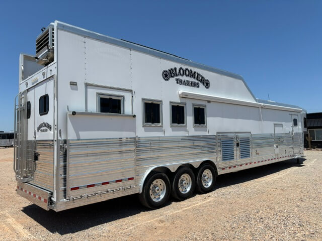 A large white Bloomer 22' loft horse trailer with multiple windows, three axles, and shiny metallic detailing is parked on a gravel lot under a clear blue sky. -Stillwater Trailer Sales